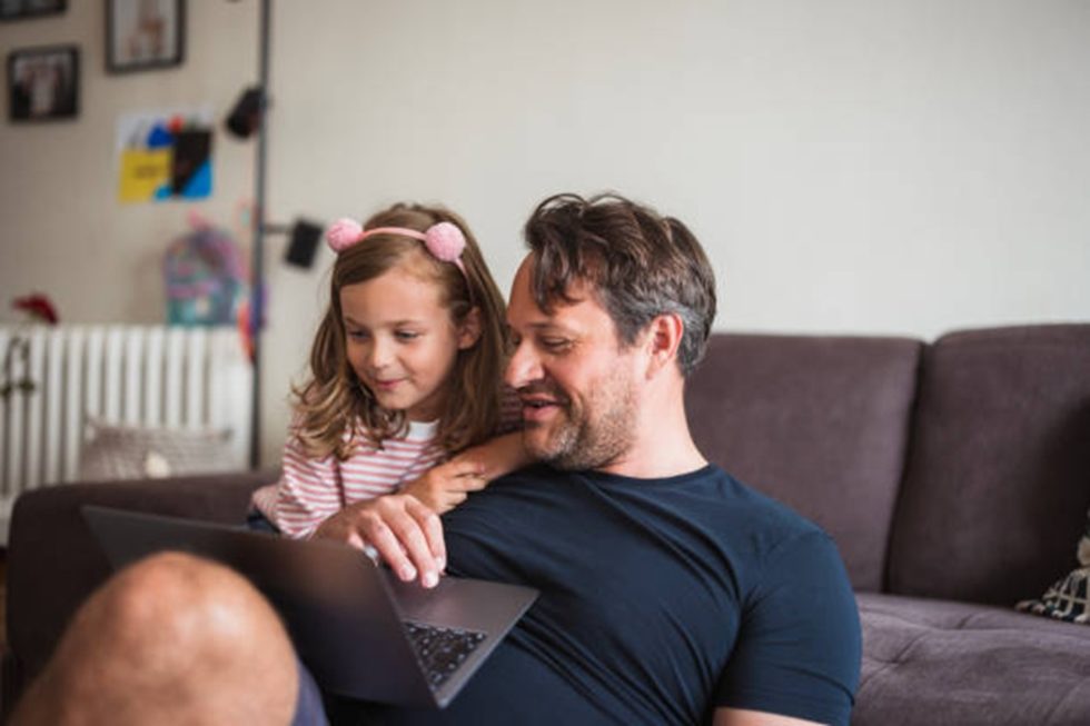 Happy young girl sitting by her dad and using laptop for watching cartoons